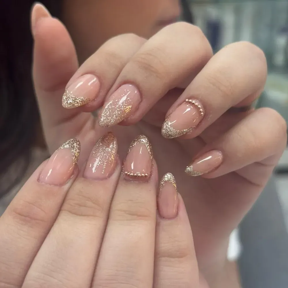 Manos con uñas en forma de almendra con esmalte nude y detalles y brillo dorado.