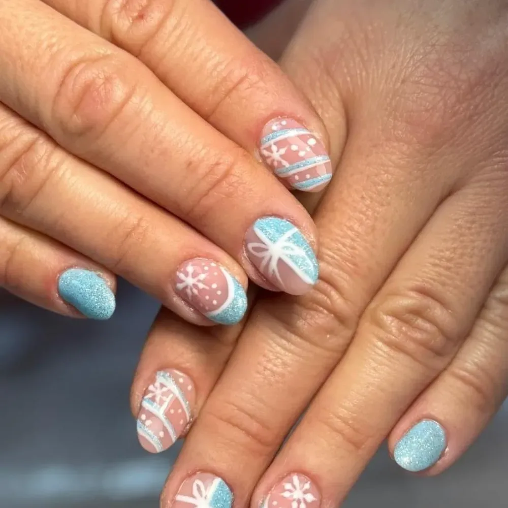 Manos con brillo celeste y nail art con temática invernal: diseños blancos sobre uñas nude con acentos celestes.