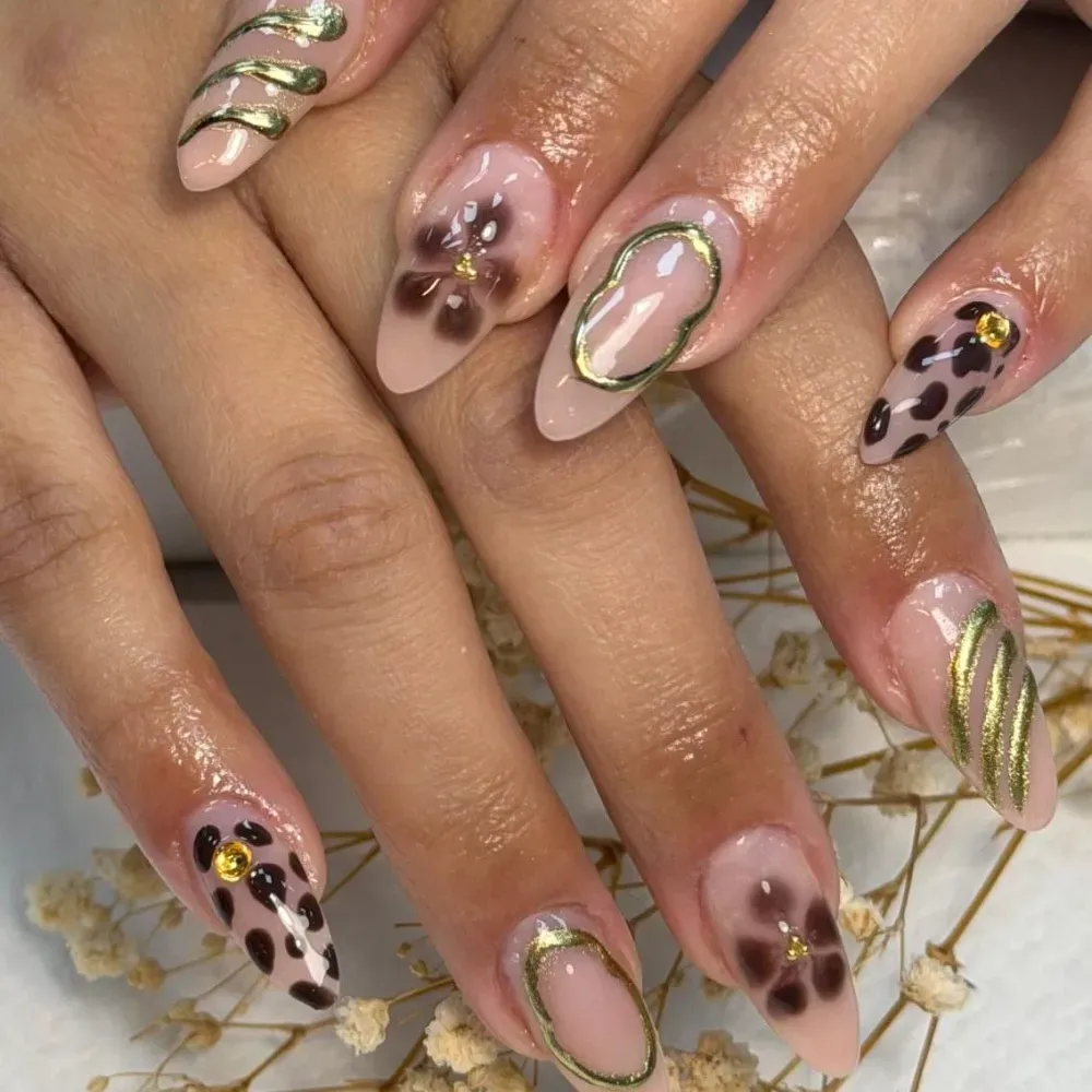 Manos con uñas en forma de almendra, con diseños florales, estampados animales y remolinos dorados sobre una base nude.