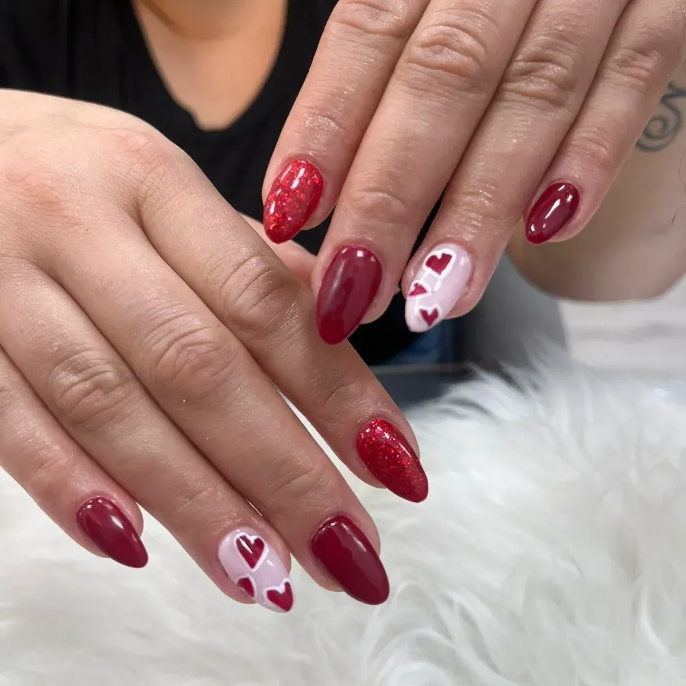 Manos con decoración de uñas de San Valentín en tonos rojos y blancos, que incluye corazones y brillantina.