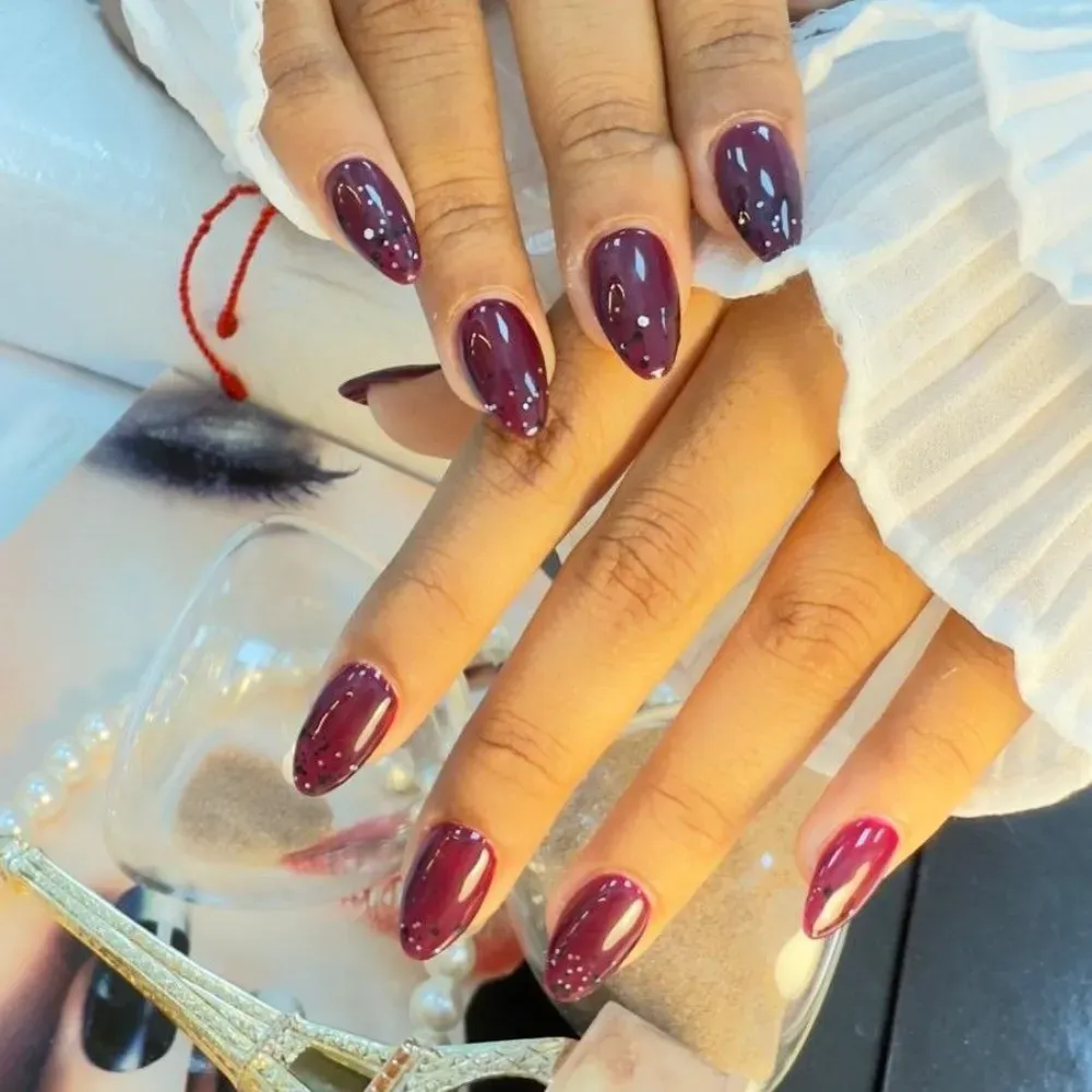 Manos con uñas ovaladas de color granate, algunas con motas de brillo blanco, que descansan sobre una superficie reflectante.