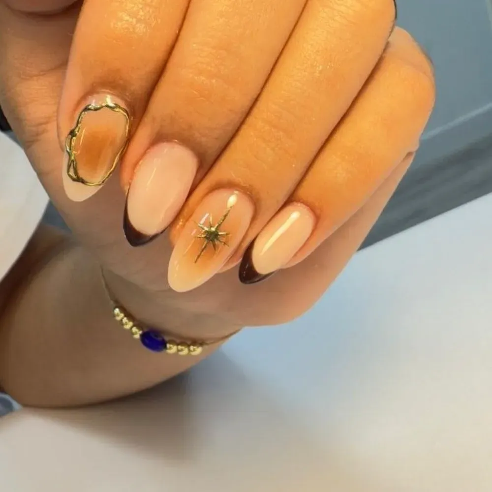 Mano con uñas almendradas, algunas con diseño dorado y detalles en marrón y negro. Se ve una pulsera dorada y azul.