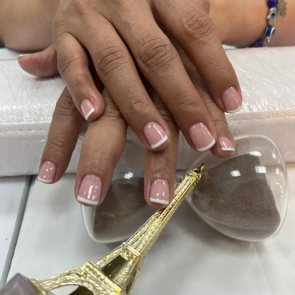 Manos con uñas de punta francesa, una Torre Eiffel dorada y un reloj de arena sobre una superficie blanca.