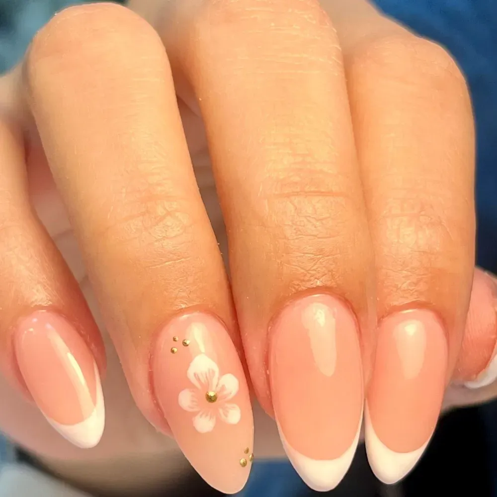 Primer plano de una mano con uñas francesas. Una uña presenta un diseño floral blanco con tachuelas doradas.