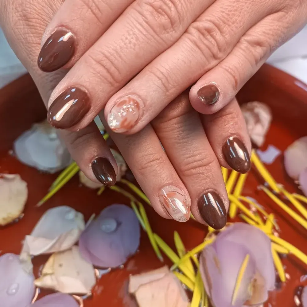 Manos con uñas ovaladas de color marrón, dos uñas con detalle de oro y perla, apoyadas sobre una bandeja roja con pétalos de flores y tallos amarillos.