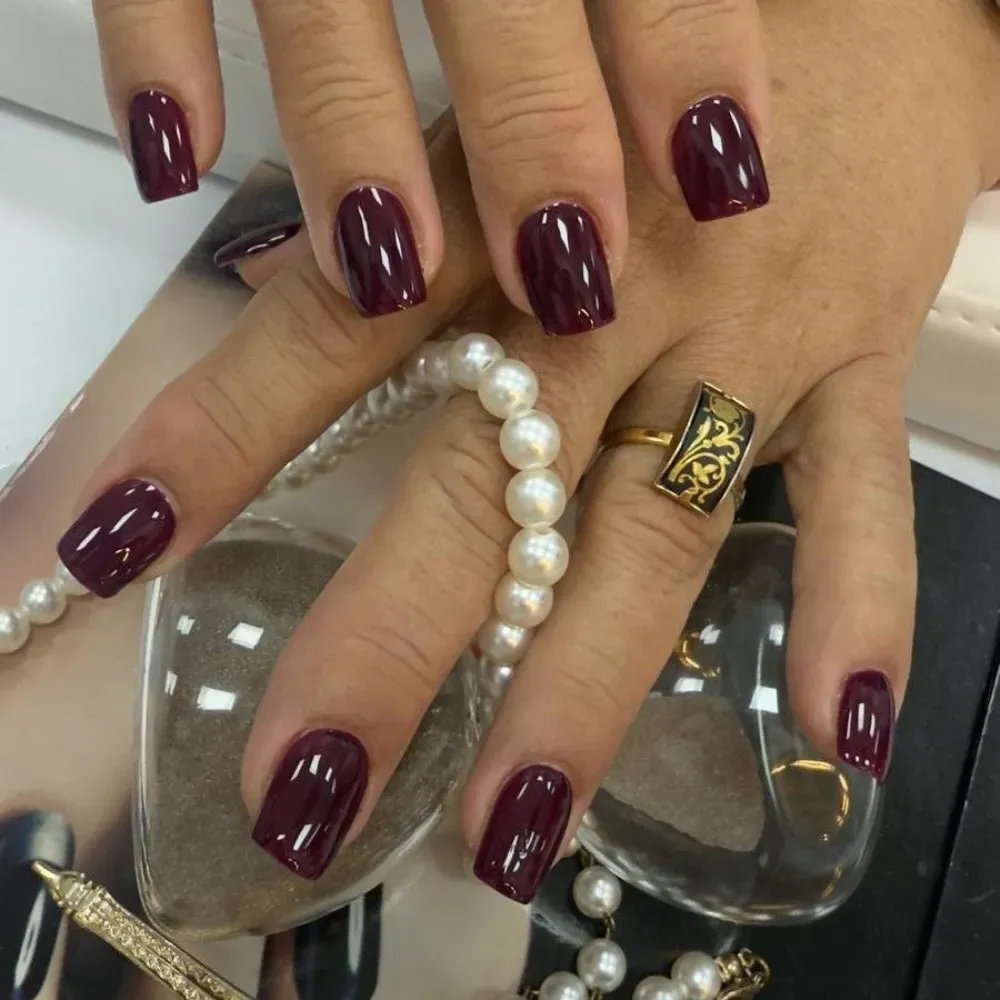 Manos de mujer con uñas cuadradas de color burdeos oscuro, anillo dorado y collar de perlas.