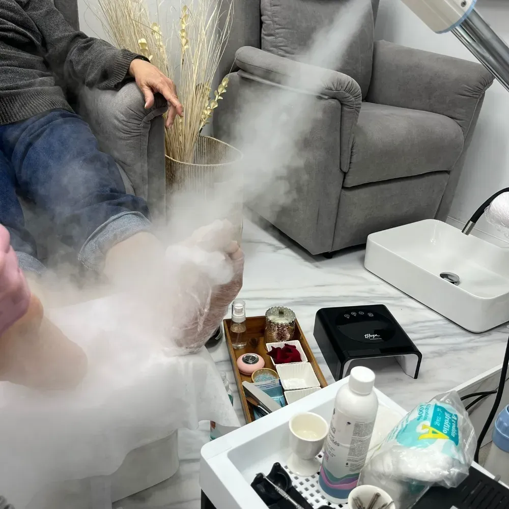 Persona recibiendo un tratamiento de vapor para pies en un salón de manicura. El vapor rodea el pie; silla gris y lavabo al fondo.
