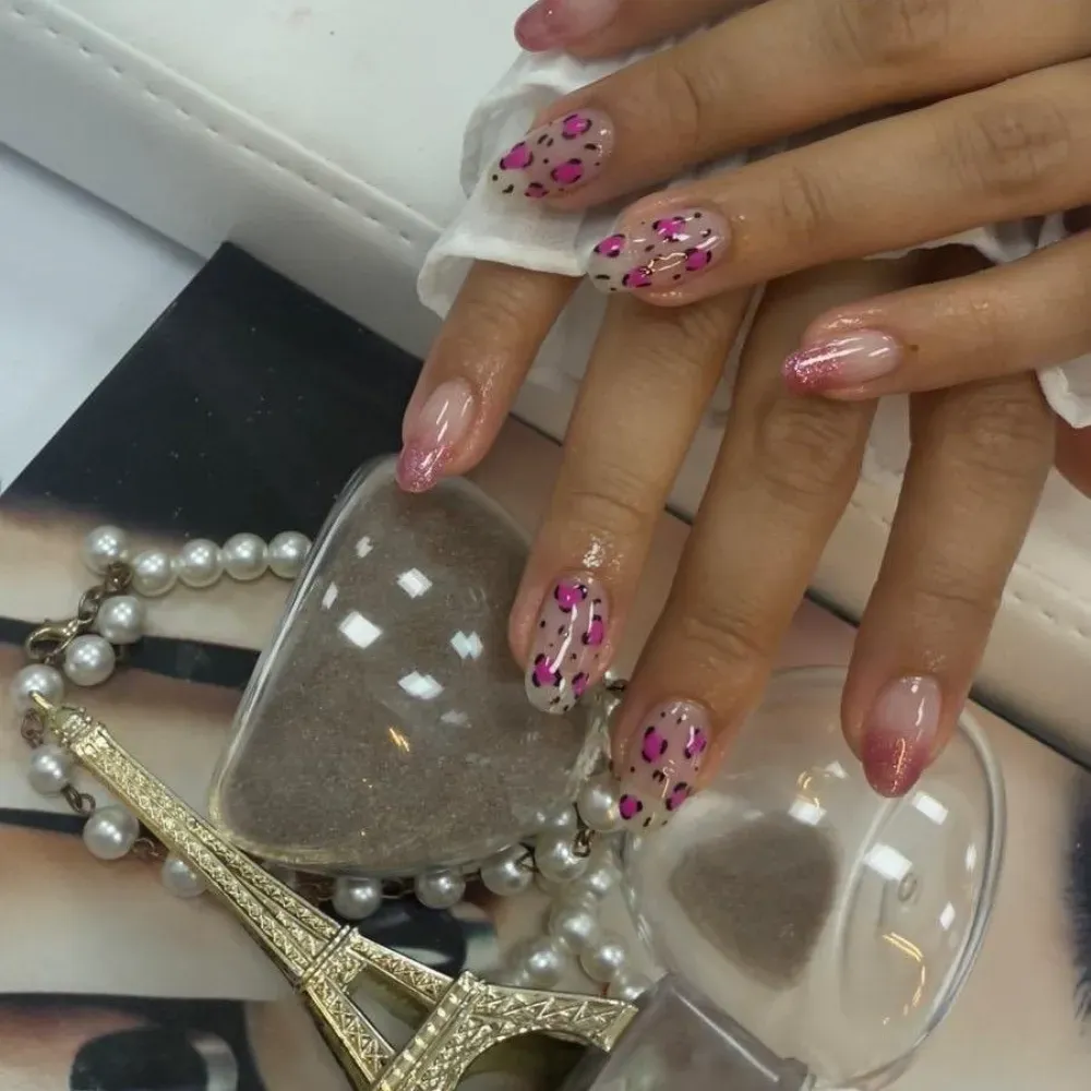 Manos de mujer con uñas rosas ombré. Una mano luce un estampado de leopardo rosa, junto con una Torre Eiffel dorada, perlas y un reloj de arena.