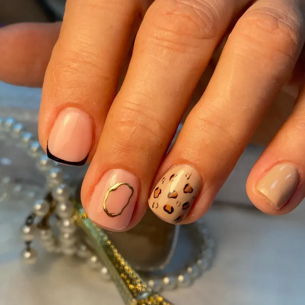 Primer plano de una mano con manicura de punta francesa y dos uñas con estampado de leopardo y contorno dorado.