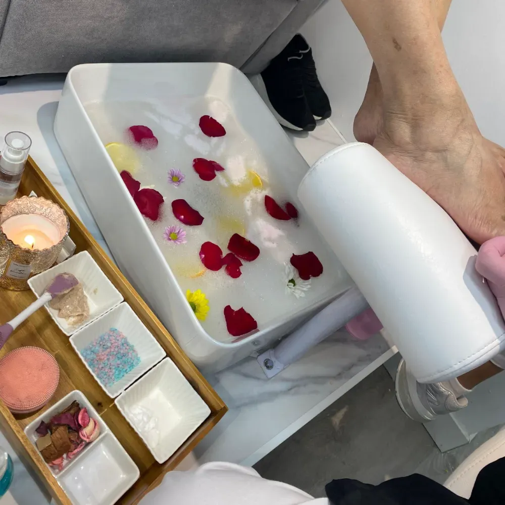 Configuración de spa para pies: pie en agua con pétalos de rosa, productos y vela en una bandeja, persona cerca.