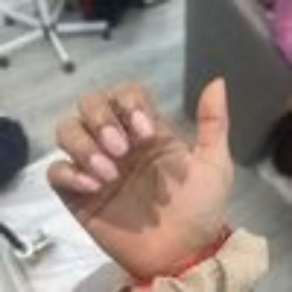 Mano con uñas ovaladas y rosadas; pulgar levantado. Muñeca con banda roja.