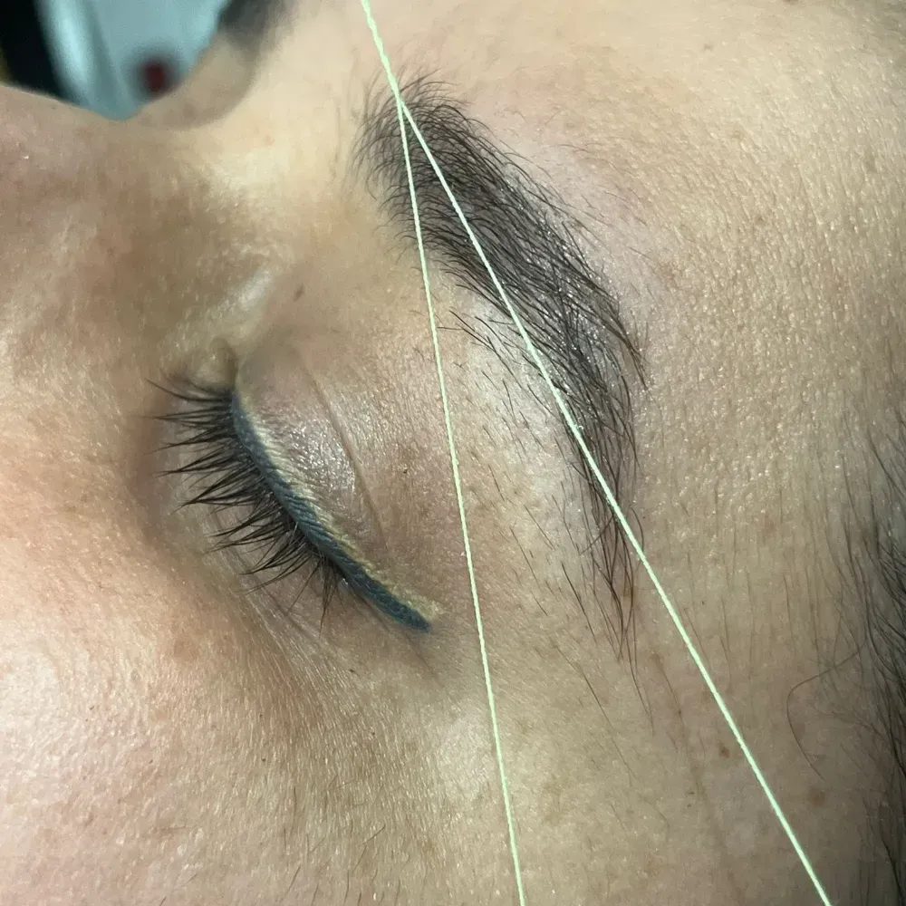 Primer plano del ojo y la ceja de una persona enhebrados con un hilo verde claro, con los ojos cerrados.