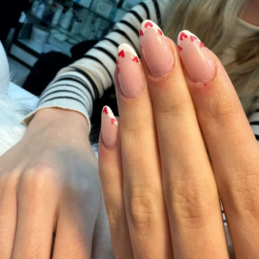 Mano de mujer con uñas largas y ovaladas. Puntas francesas con pequeños corazones rojos adornan las uñas.