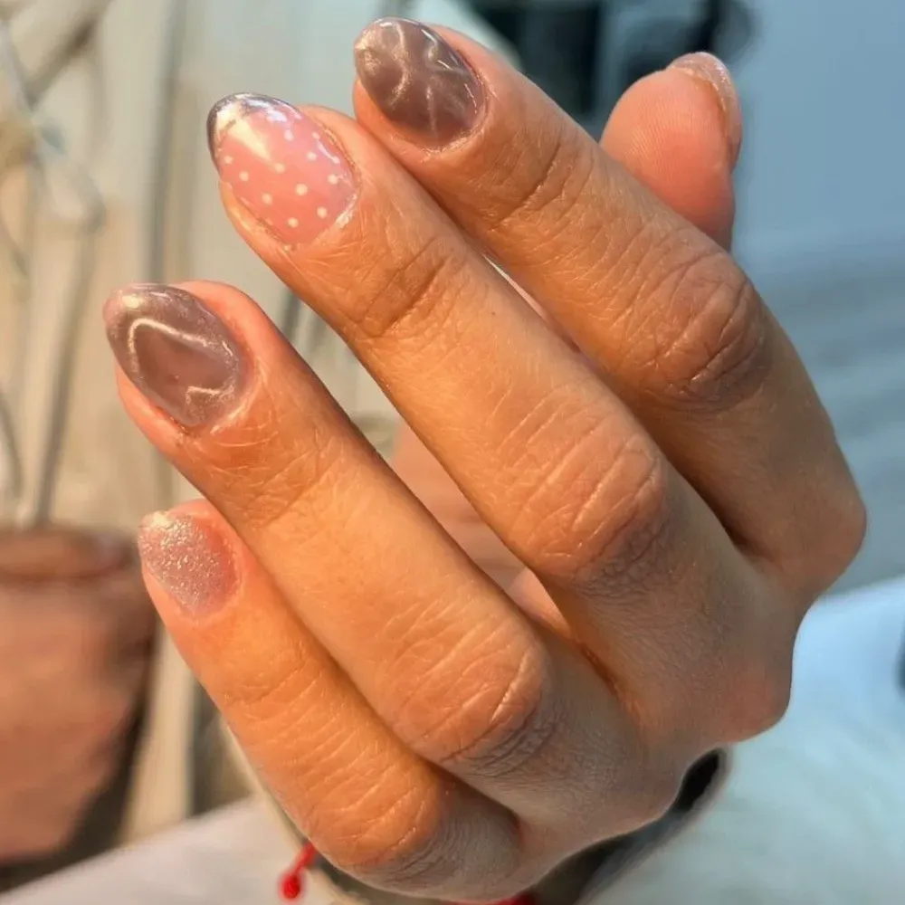 Primer plano de una mano con uñas de color marrón claro, algunas con detalles plateados o rosas, una con lunares blancos.