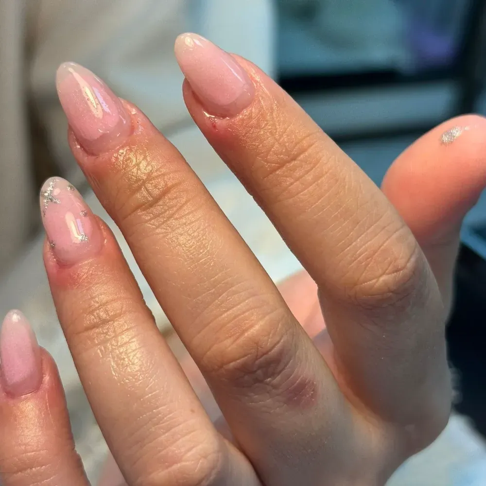 Mano con uñas largas, brillantes y de color rosa claro; una de las uña tiene brillo plateado.