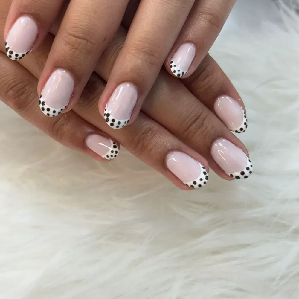 Primer plano de manos con uñas de color rosa claro, cada una con una punta blanca decorada con pequeños puntos negros.