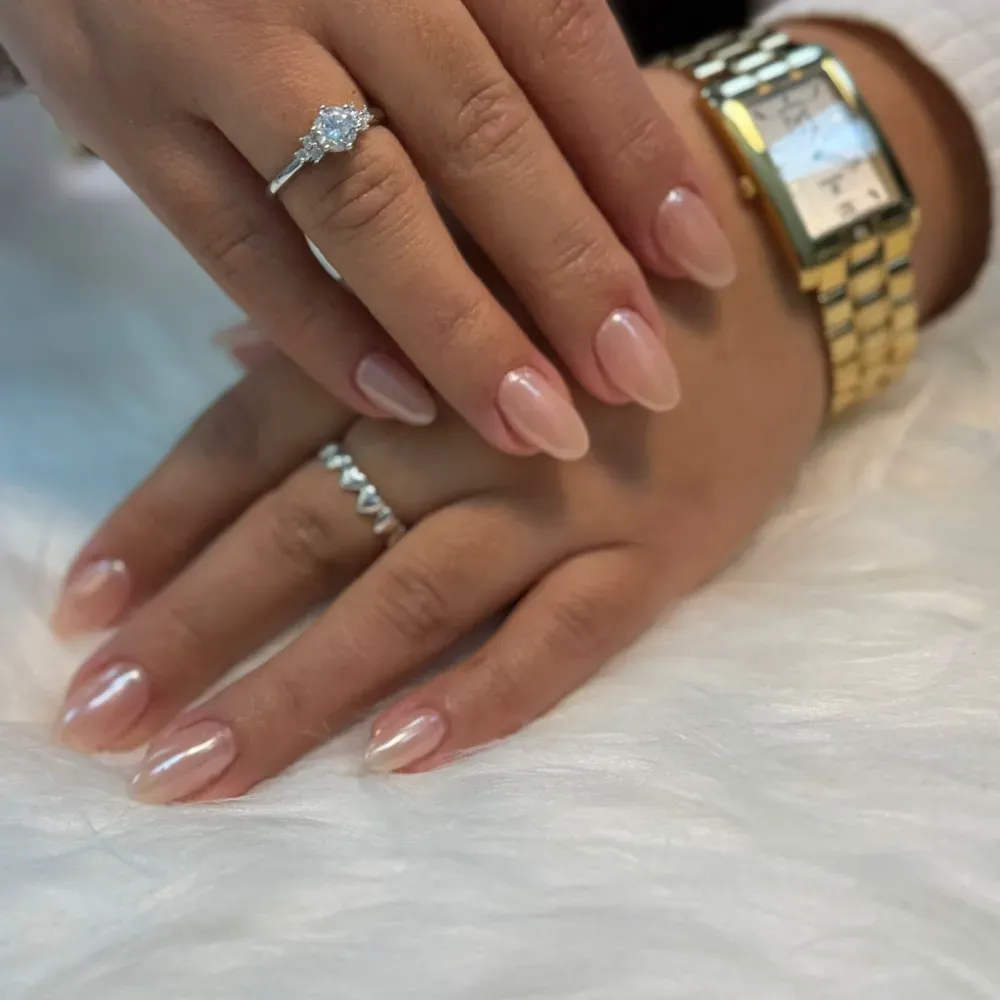 Manos con uñas ovaladas color nude, anillos y un reloj dorado sobre una superficie blanca y esponjosa.
