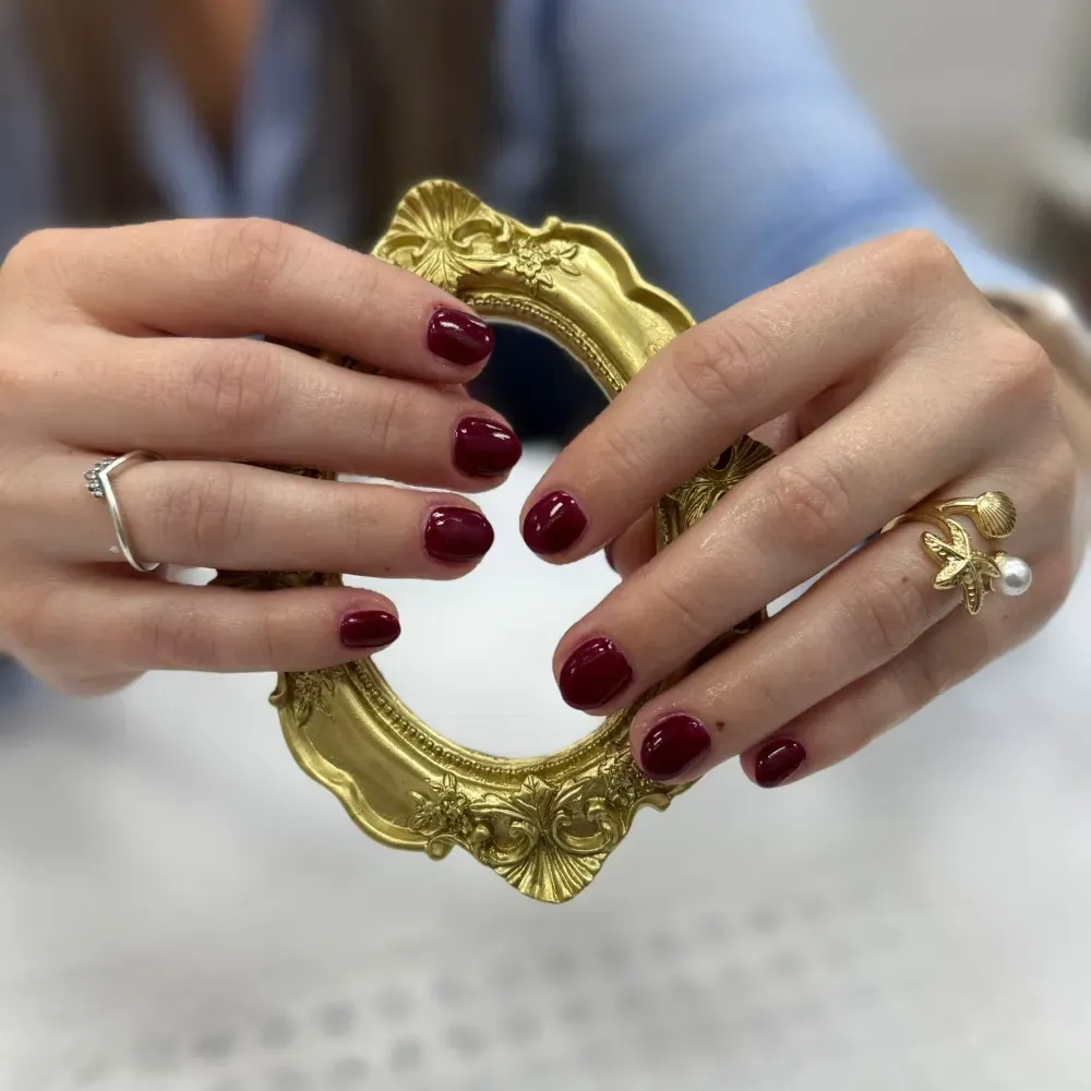 Manos con esmalte de uñas de color rojo oscuro sosteniendo un marco dorado adornado, con anillos en los dedos.