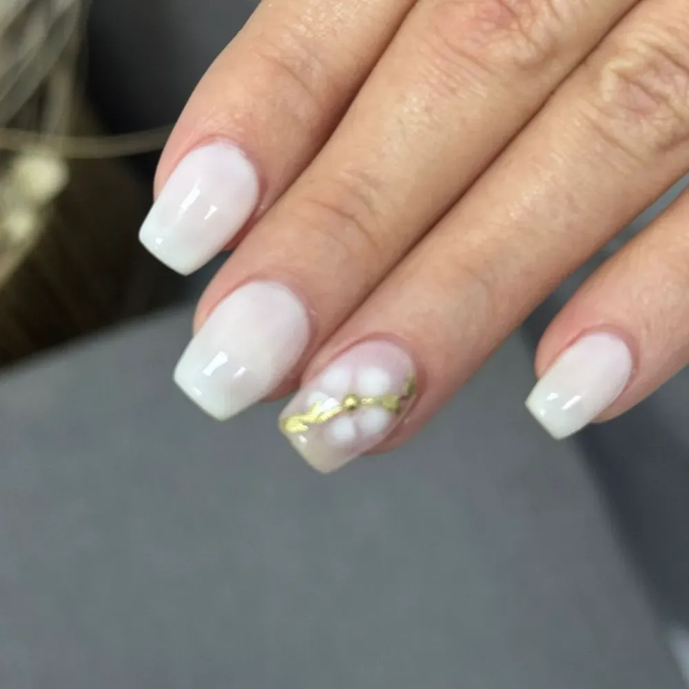 Mano de mujer con uñas blancas cuadradas. Una de ellas luce un diseño floral dorado.