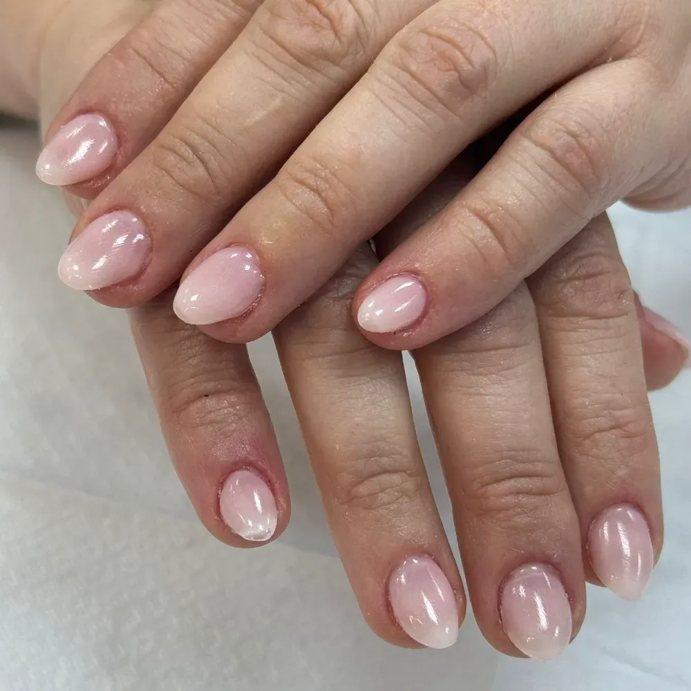 Manos con uñas de forma ovalada, pintadas en un degradado de rosa claro a blanco, apoyadas sobre una superficie blanca.