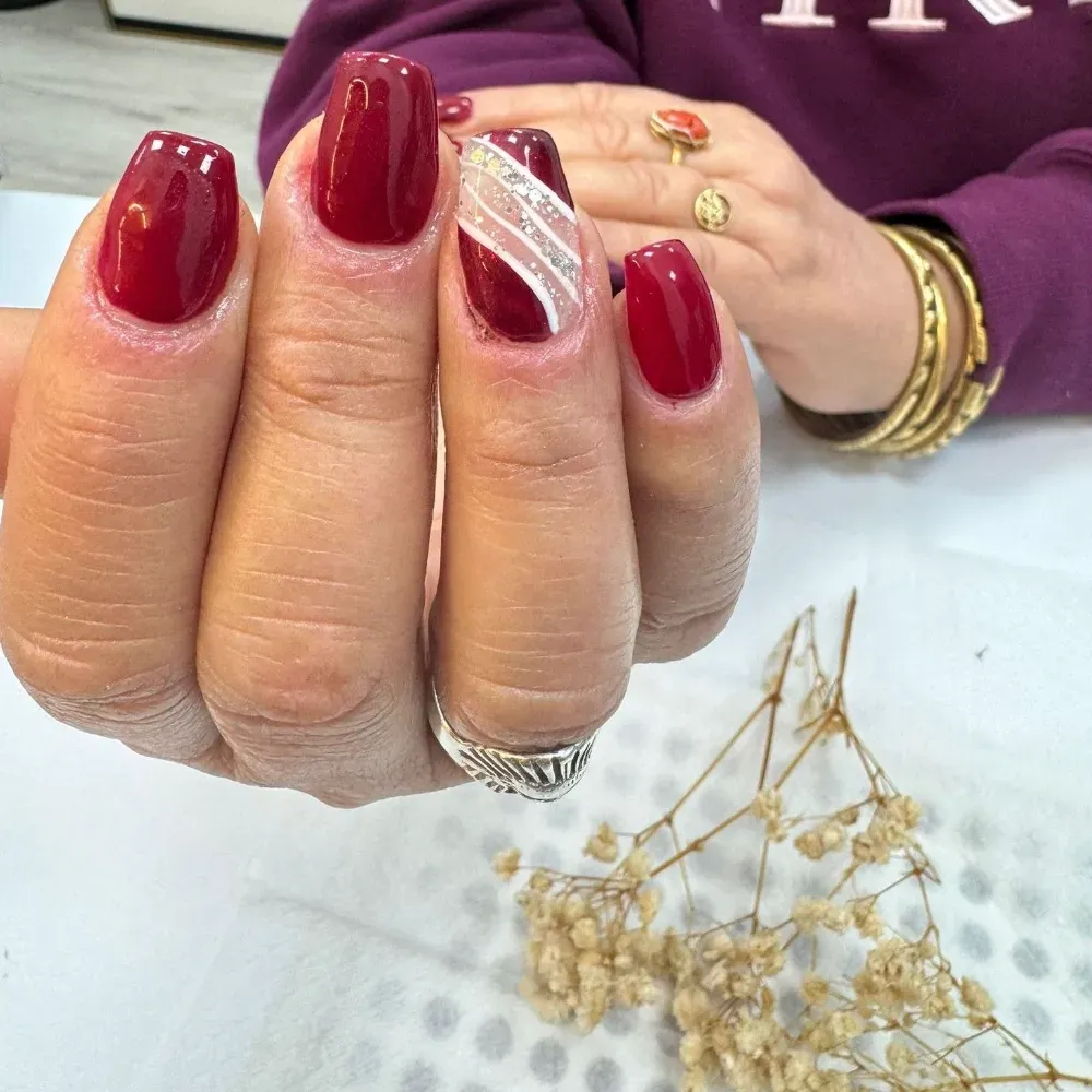 Mano con uñas rojas en forma de ataúd, una de ellas con diseño de remolino blanco; anillos y pulseras de oro, ramita de flores secas.