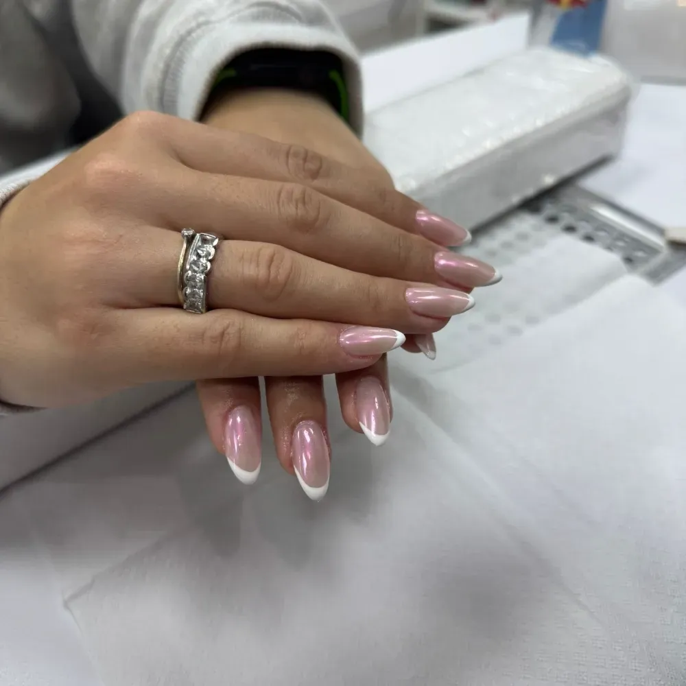 Mano con diseño de uñas con punta francesa, anillo, apoyada sobre una superficie blanca, con suministros de manicura en el fondo.