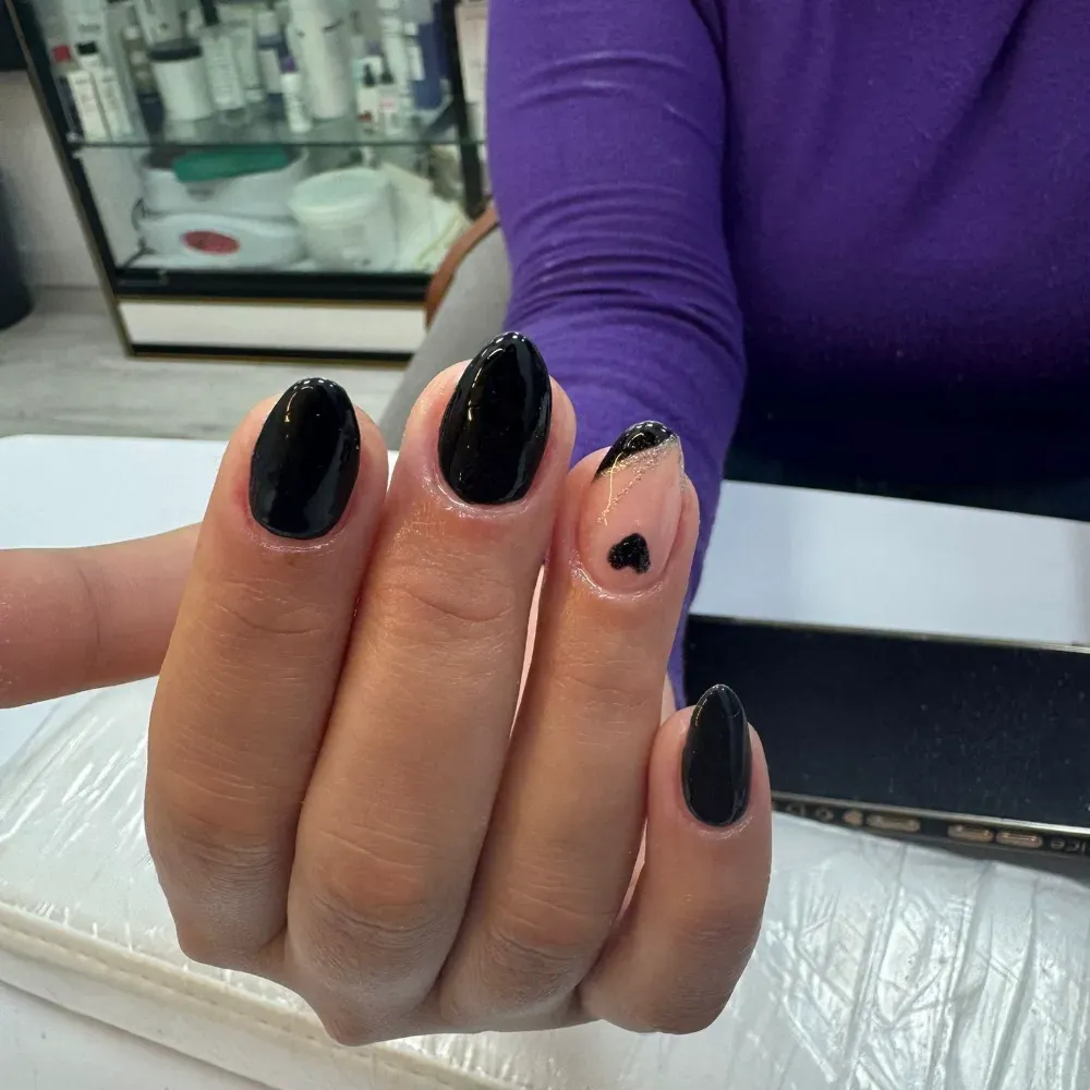 Mano con uñas negras ovaladas, una con un diseño rosa y un pequeño corazón. En un salón de manicura.