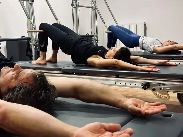 Personas practicando Pilates en máquinas reformer en un estudio, tumbadas sobre colchonetas y elevando las caderas.