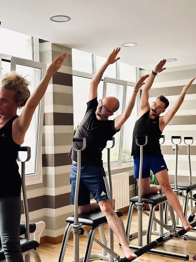 Personas haciendo ejercicio en máquinas de Pilates en un estudio luminoso, estirando los brazos por encima de la cabeza.