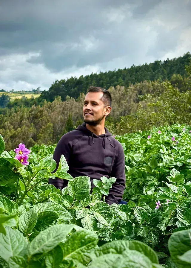 Un hombre de pie en un campo verde exuberante con flores moradas bajo un cielo nublado.