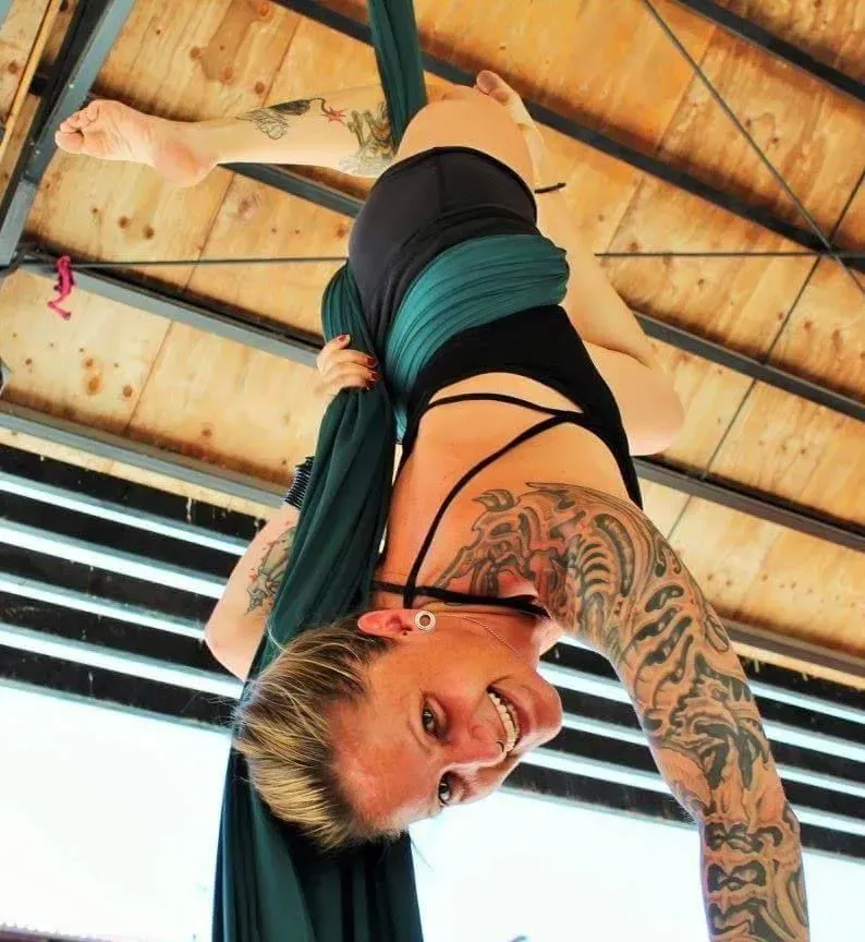 Mujer colgando boca abajo de telas aéreas bajo un techo de madera, sonriendo con el brazo tatuado visible.