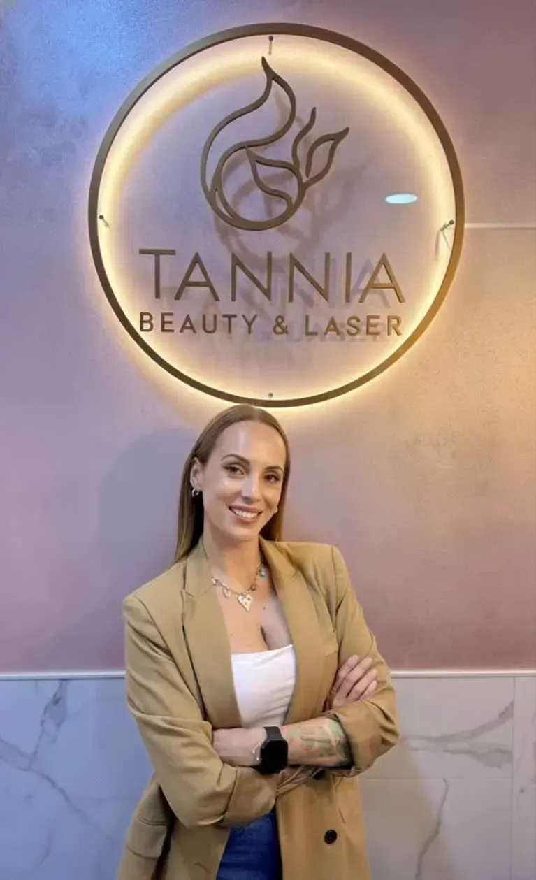 Mujer con los brazos cruzados sonríe, vistiendo un blazer color canela, una blusa blanca y joyas; en el interior.