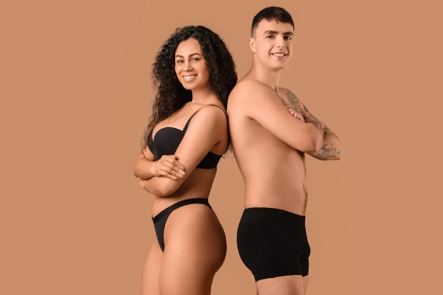 Mujer en sujetador negro y hombre con tatuajes, ambos sonriendo, con los brazos cruzados.