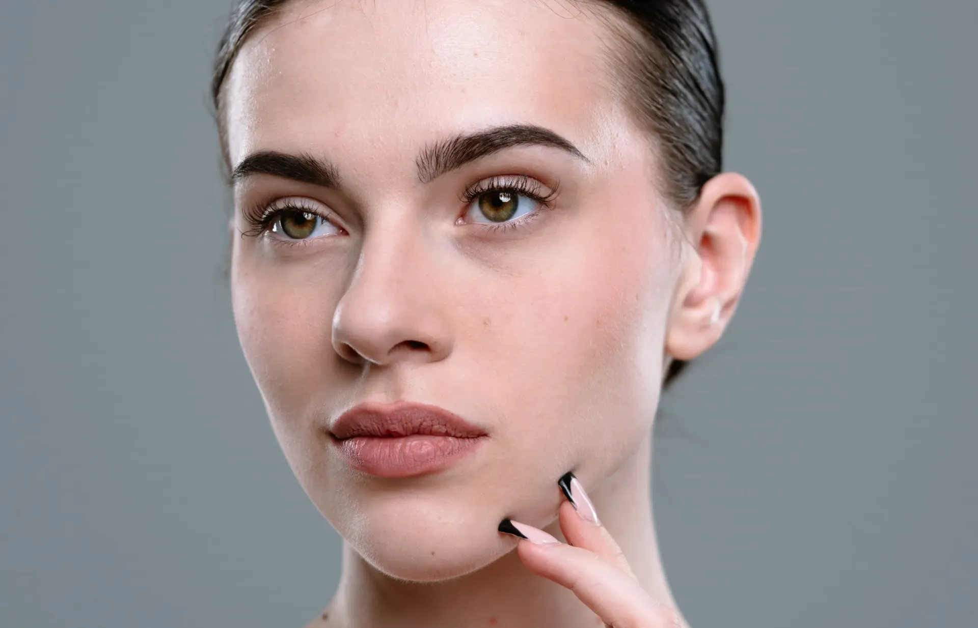 Mujer de cabello oscuro, mirando hacia un lado, tocándose la cara. Fondo gris.