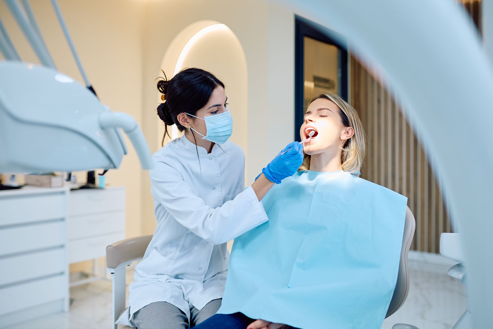 Un dentiste examine la bouche d'un patient dans une clinique dentaire lumineuse, avec une bavette bleue et des instruments visibles.