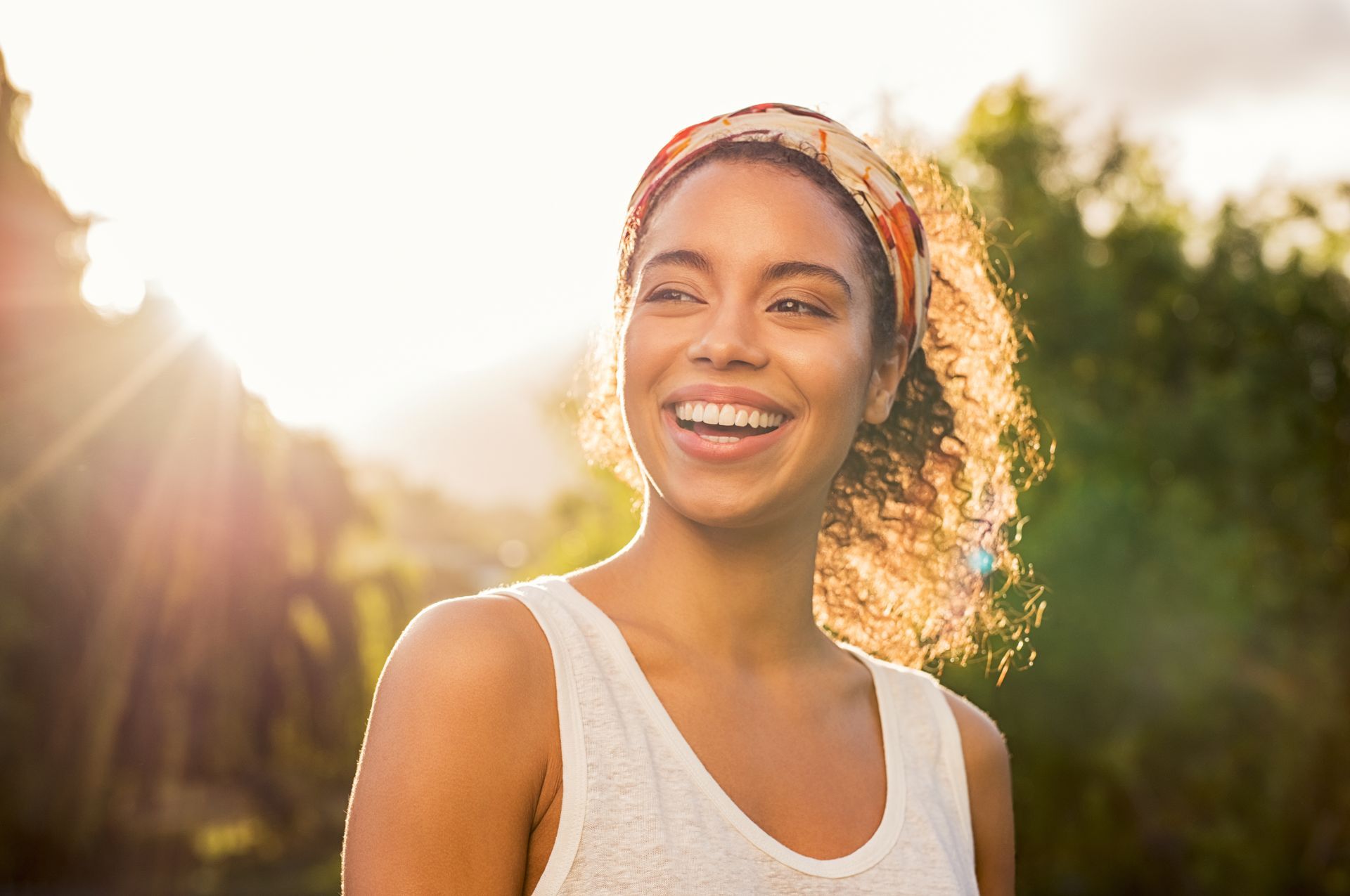 Personne souriante à l'extérieur, sous un soleil chaud, portant un débardeur blanc et un bandeau à motifs