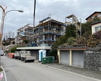 Ein im Bau befindliches Gebäude mit Gerüst, einem Kran und geparkten Lastwagen. Garagen und eine Strasse im Vordergrund.