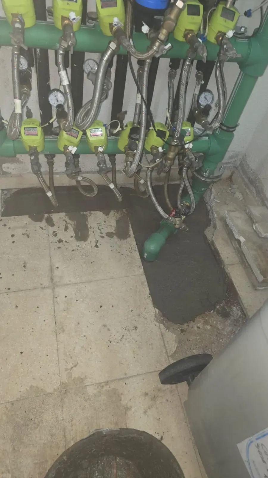 Maquinaria verde con cables y mangueras colgando sobre un suelo de baldosas mojado y sucio en una sala industrial.