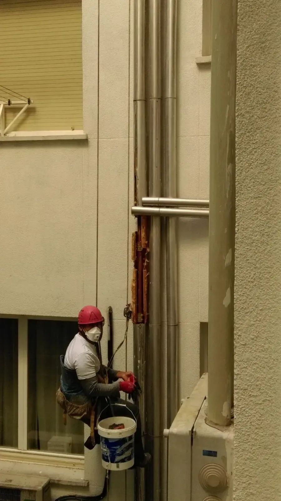 Obrero con casco rojo pintando tuberías altas en la pared de un edificio industrial.