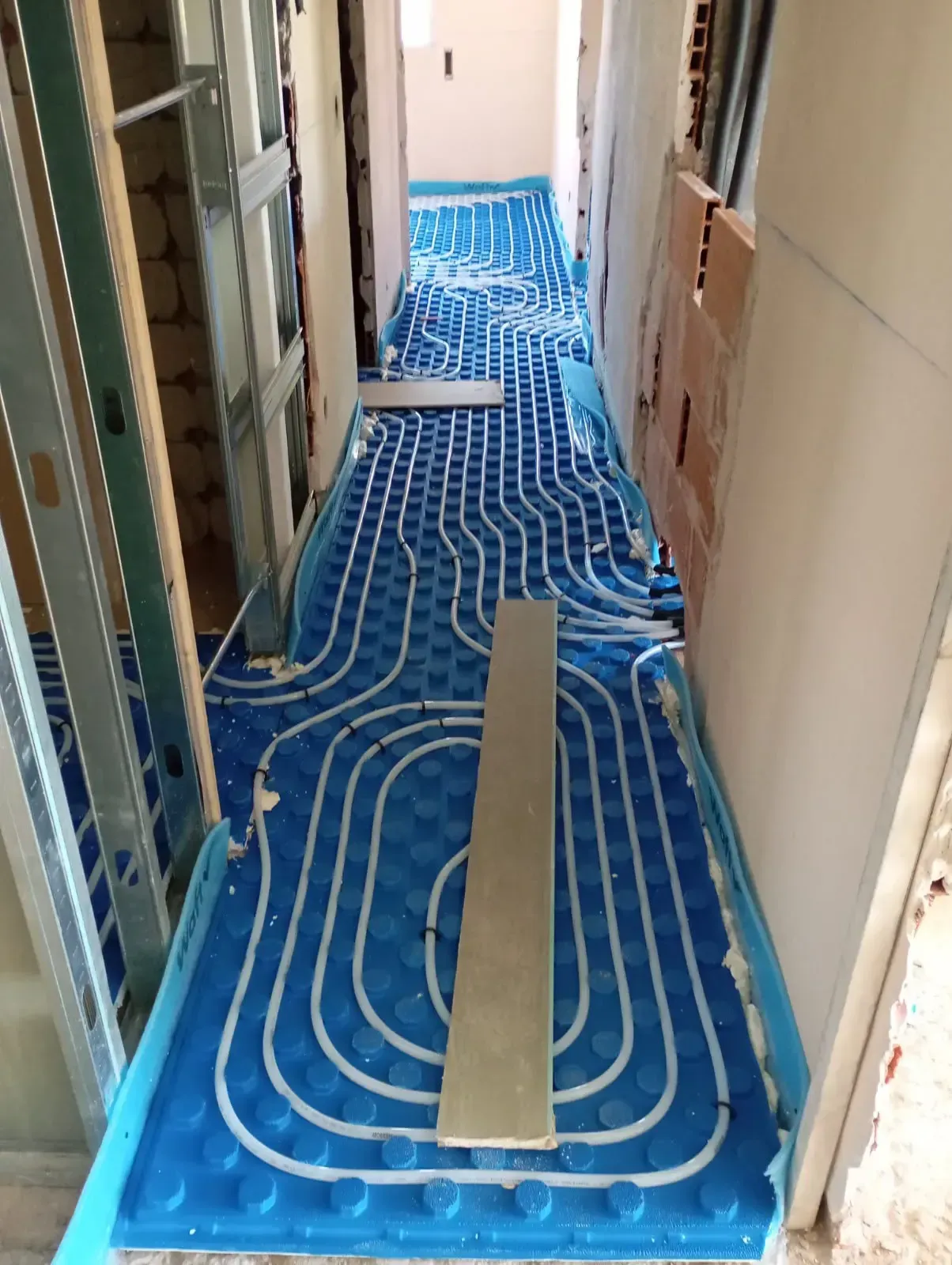 Pasillo en obras con tuberías azules de calefacción por suelo radiante tendidas a lo largo del suelo y paredes sin terminar.