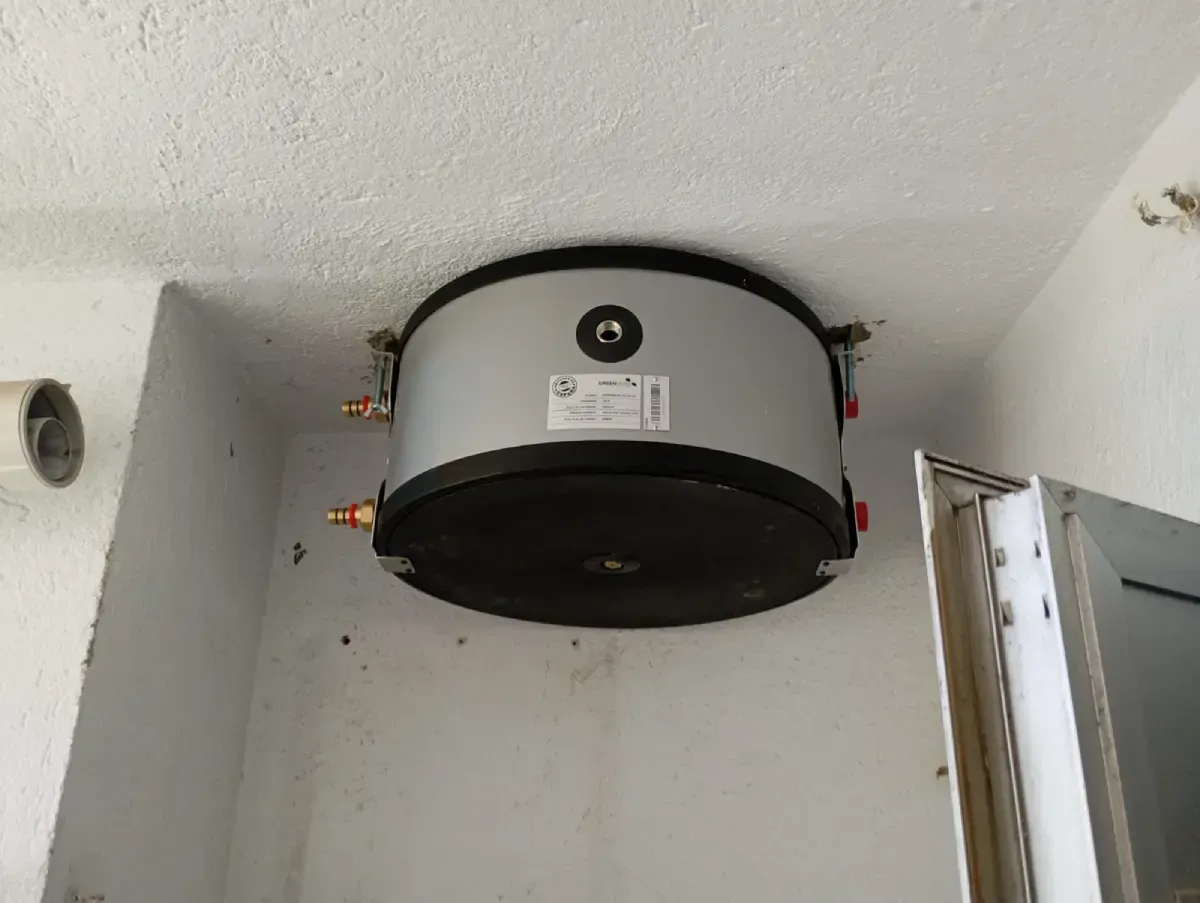 Tanque cilíndrico negro montado en el techo de un cuarto de servicio, con tuberías y válvulas en una pared blanca.