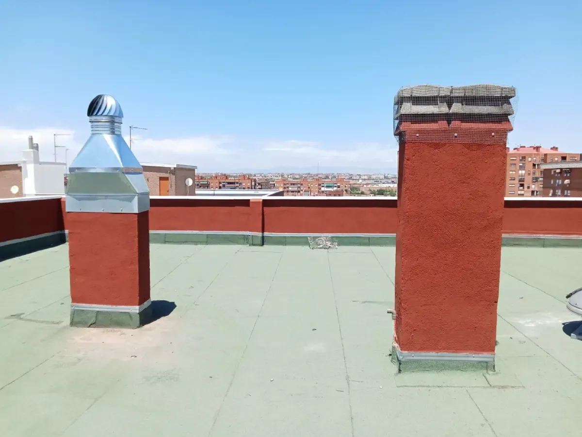 Azotea con superficie verde, chimeneas de ladrillo rojo y una rejilla de ventilación plateada bajo un cielo azul.