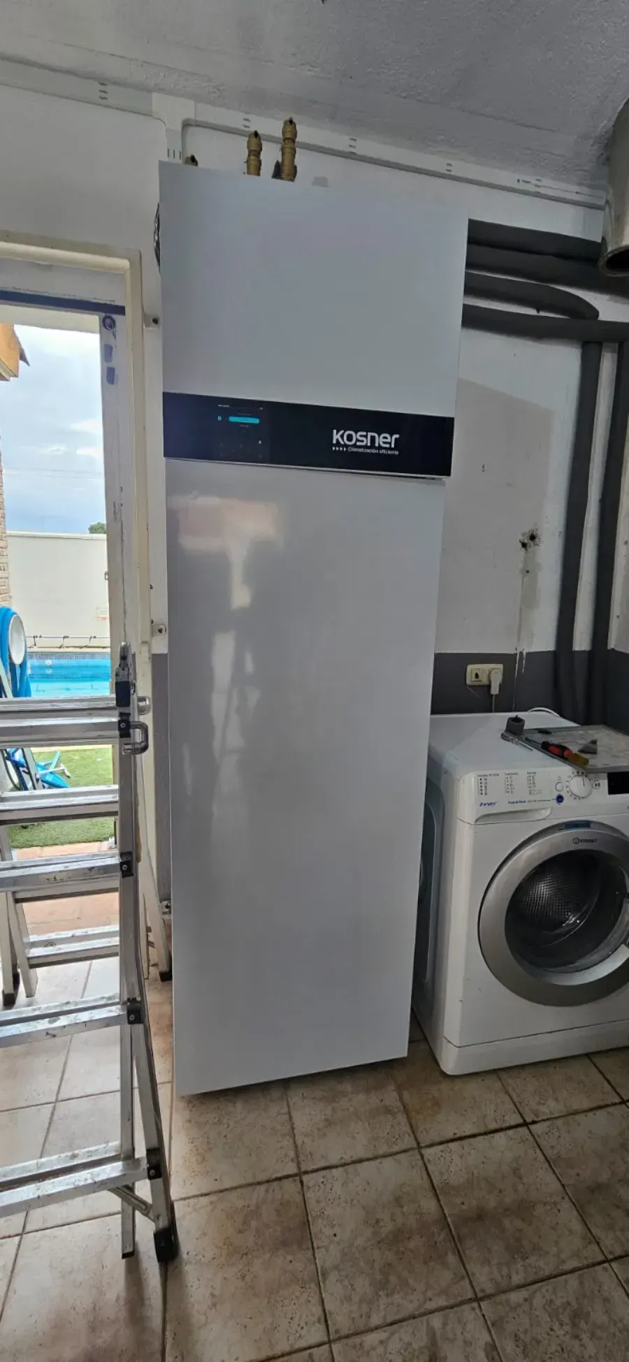 Refrigerador blanco en un cuarto de lavado junto a una lavadora, con piso de baldosas y tuberías de servicios