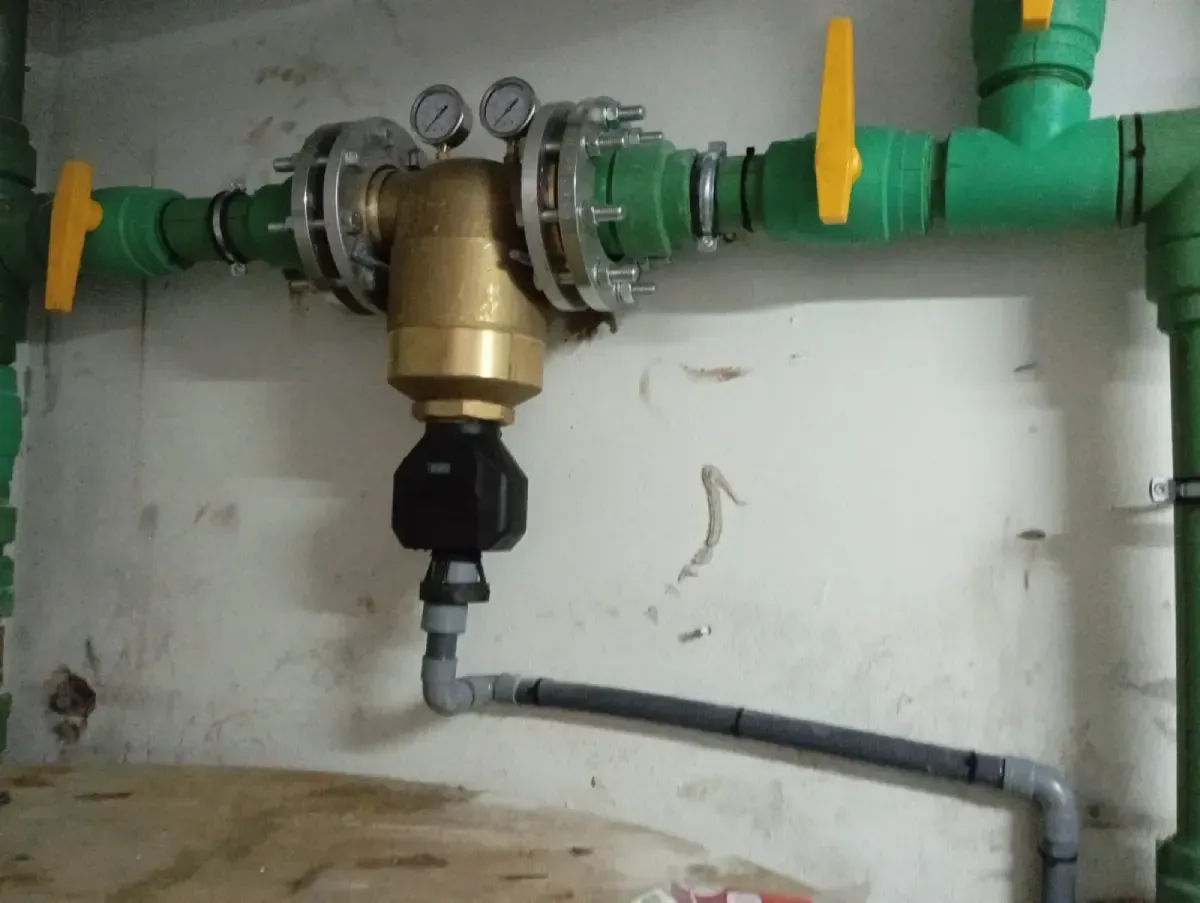 Tuberías de fontanería con regulador de presión de latón, manómetros y válvula de cierre amarilla contra una pared blanca.
