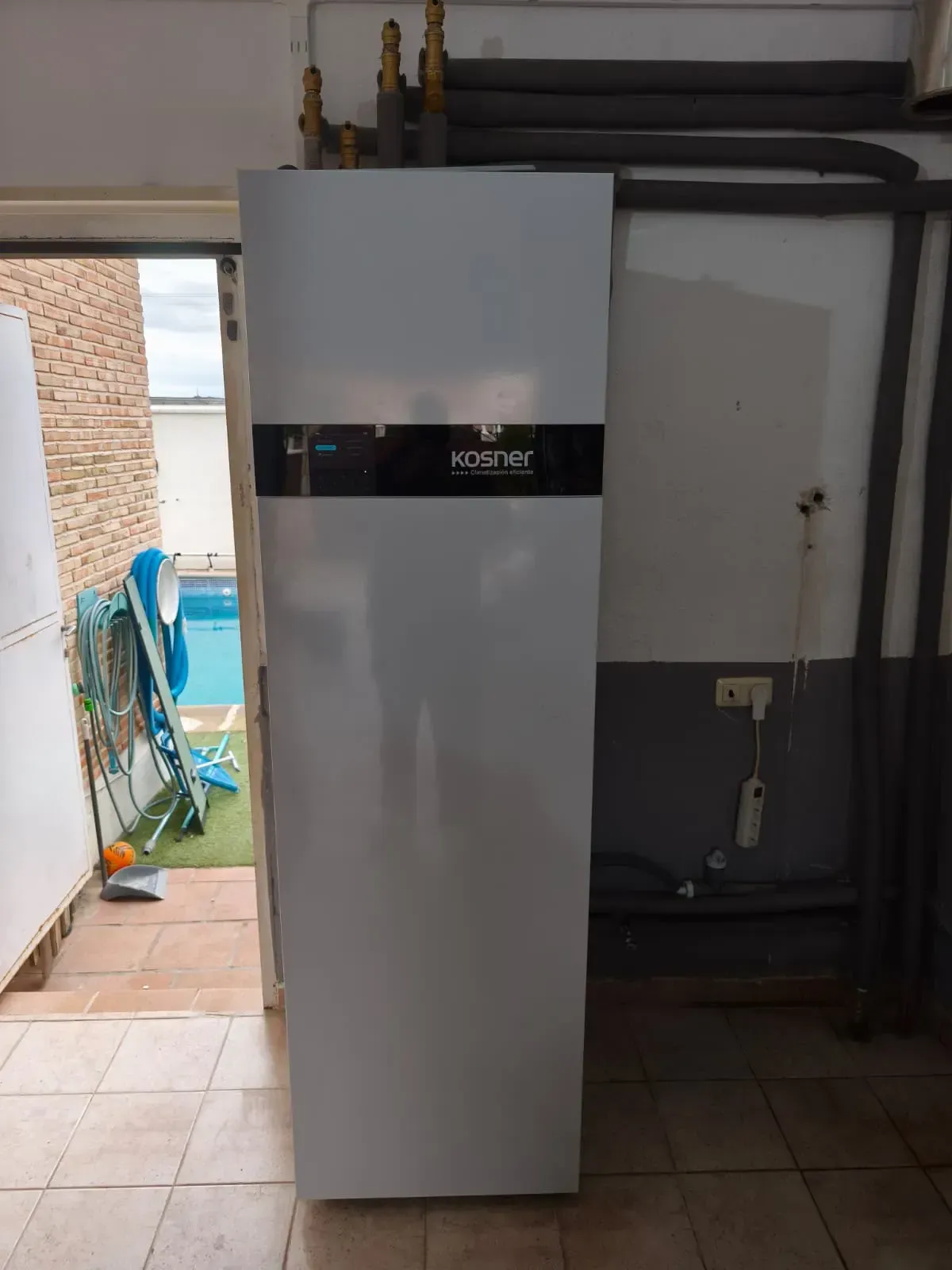 Electrodoméstico blanco alto con panel central negro en un cuarto de servicio con azulejos; puerta y tuberías al fondo.