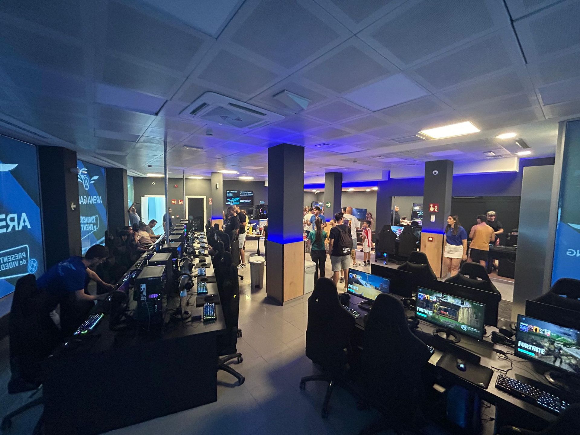 Centro de juegos lleno de gente jugando con computadoras. Iluminación azul y un ambiente moderno.