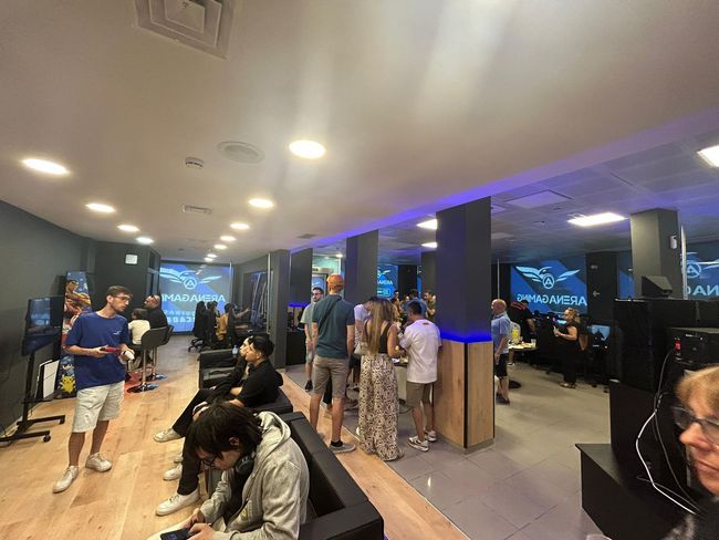 La gente se reunía en una sala de juegos, algunos sentados frente a las computadoras, otros socializando. Iluminación tenue, detalles en azul.