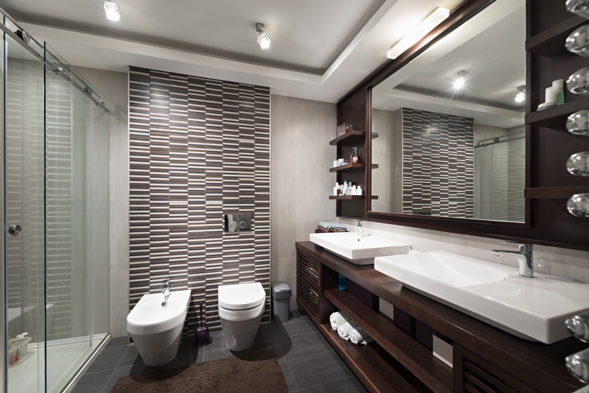 Salle de bains moderne avec douche en verre, deux toilettes, grand miroir et meuble-lavabo en bois foncé.