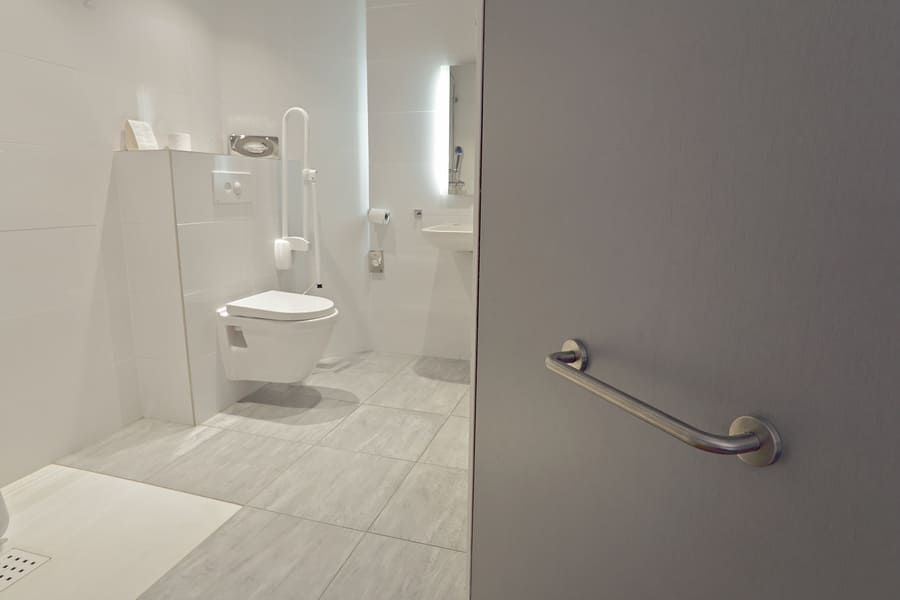 Salle de bain accessible avec toilettes, barres d'appui et porte ouverte dans un intérieur blanc lumineux