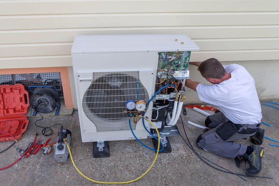 Un technicien répare une unité de climatisation extérieure à côté d'une maison, avec ses outils et ses câbles au sol.